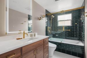 Modern bathroom with a wood vanity, white countertop, gold fixtures, black subway tile in the shower, glass shower door, and a backlit mirror above the sink.
