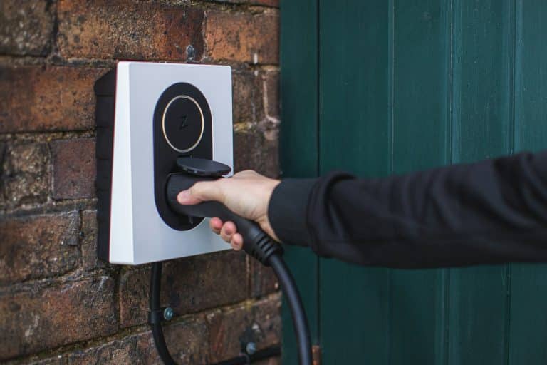 A person plugs a charging cable into an electric vehicle charger mounted on a brick wall next to a green door, illustrating how EV charging at home can offer convenience while future-proofing your lifestyle.