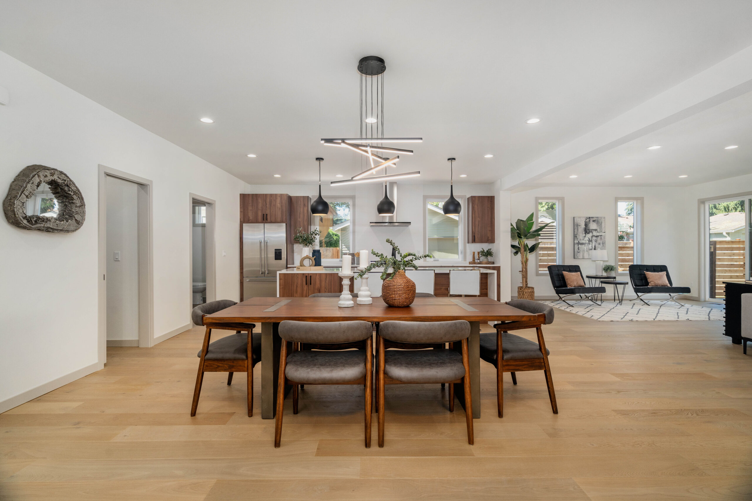 A7309636 Modern open-concept kitchen and dining area with a wooden table, six chairs, pendant lights, and stylish decor. Living area with large windows and neutral tones visible in the background.