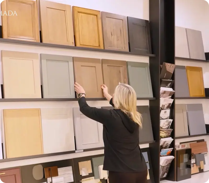 A woman in a black shirt reaches for a wooden cabinet door sample on a display wall filled with various colors and finishes.
